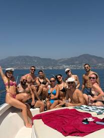 A lively group enjoys a fun day on the water, celebrating with drinks and sunbathing aboard a boat, surrounded by stunning coastal views.
