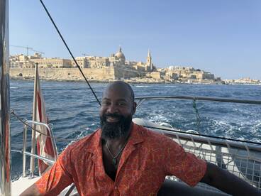 A smiling man enjoys a scenic boat ride with a stunning cityscape in the background, perfect for capturing memorable vacation moments.
