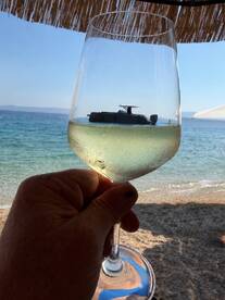 A hand holding a glass of wine with a boat reflected in it, set against a beach backdrop. The unique perspective creates an artistic view of a relaxing day by the water.
