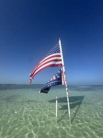 A flagpole displaying an American flag and another flag in shallow water, with a clear blue sky above. The scene captures a serene coastal atmosphere.