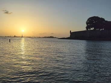 A stunning sunset view over the harbor, featuring a silhouette of a historic fort and a sailboat in the distance — perfect for capturing the beauty of coastal destinations.