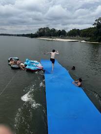 A lively scene of friends enjoying a day on the water, with one jumping off a floating platform while others relax on inflatable loungers — perfect for summer fun and water activities.
