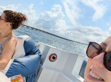 Two friends enjoy a sunny day on the water, capturing the fun of their boating experience with a picturesque pier in the background.