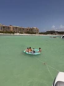 A lively group enjoys a fun day on a floating mat in the clear waters, surrounded by a beautiful beach backdrop — perfect for summer adventures.
