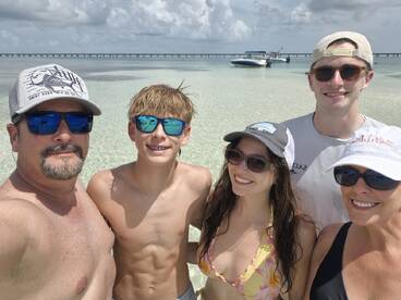 A cheerful family poses for a selfie in shallow waters, with a boat anchored in the background — capturing a perfect day at the beach.