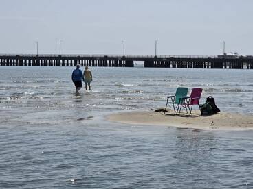 A couple strolls hand-in-hand through shallow waters at the beach, with a pier in the background, creating a serene coastal scene perfect for romantic getaways.