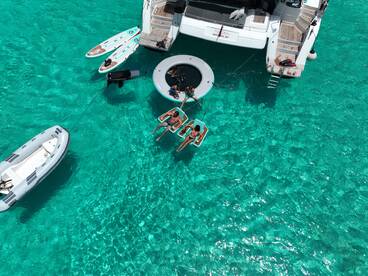 A lively group enjoys a day on the water, relaxing on floating mats and paddleboards near a luxurious yacht in stunning turquoise waters.