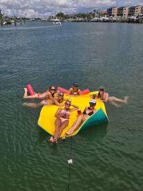 A fun group of friends relaxes on a floating mat in the water, enjoying drinks and sunshine — perfect for a summer day on the water.
