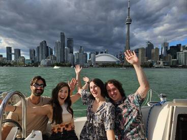 A cheerful group of friends waves from their boat, with the iconic Toronto skyline in the background — perfect for memorable city tours on the water.