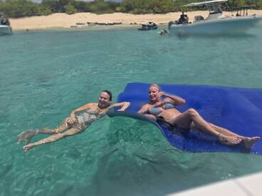 Two women enjoy a relaxing day in the clear waters, lounging on a floating mat near a beach — a perfect scene for summer fun and relaxation.