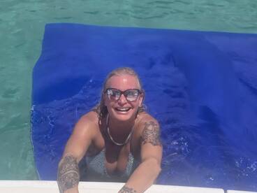 A joyful woman enjoys a sunny day swimming and relaxing on a floating mat in crystal-clear waters, perfect for a fun day at the beach or on a boat.