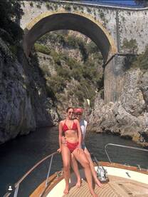 A couple enjoys a sunny day on a boat, posing against a stunning rocky backdrop and arching bridge — a perfect snapshot of summer adventures on the water.