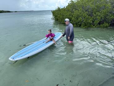 A child enjoys paddleboarding with an adult in shallow waters surrounded by mangroves, showcasing a fun and engaging water activity perfect for family outings.