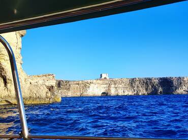 A picturesque view of a rugged coastline and a historic landmark from a boat, showcasing the stunning blue waters and clear skies — perfect for scenic boat tours.
