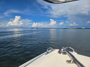 A serene view from the bow of a boat, showcasing a picturesque bridge under a bright blue sky with fluffy clouds — perfect for capturing the tranquility of a day on the water.
