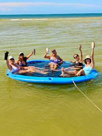 A lively group of friends enjoys a fun day on a floating mat, raising their drinks in celebration under the sun — perfect for summer getaways and beach parties.