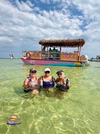 Three friends enjoy a refreshing drink while wading in the clear waters next to a vibrant tiki boat in Destin, Florida — a perfect setting for a fun day on the water.