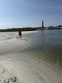 A woman walks her dog along a sandy beach with a picturesque lighthouse in the background, capturing a serene moment in nature.