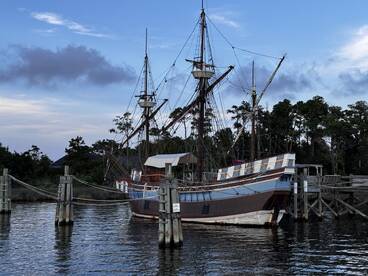A beautifully detailed ship docked at the pier, showcasing its impressive sails and hull against a serene backdrop of trees and sky.