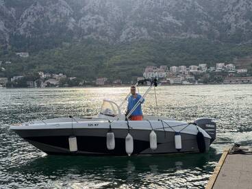 A man gives a thumbs up while standing on a boat in serene waters, with picturesque mountains and a charming village in the background — a perfect snapshot of a boating adventure.