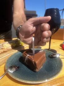 A hand holds a birthday cake with a candle, accompanied by a glass of wine, perfect for celebrating special moments on a boat.