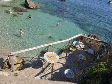 A vibrant scene of people swimming in crystal-clear waters, with a picturesque beachside setting featuring colorful tables and chairs — perfect for enjoying a sunny day by the sea.