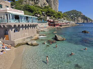 A vibrant beach scene with people enjoying the clear waters and sunbathing near a picturesque restaurant along the coastline, perfect for a relaxing day out.