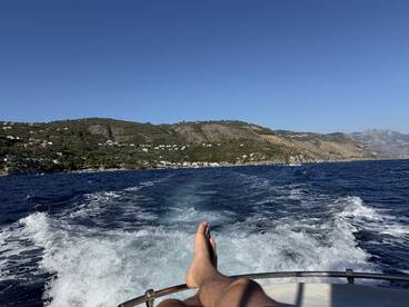 A scenic view from a boat with a person's foot resting on the edge, showcasing the beautiful coastline and clear blue waters — perfect for a relaxing day on the water.
