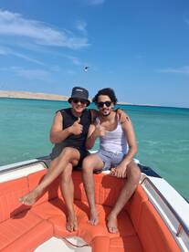 Two friends enjoy a sunny day on a boat, giving thumbs up against a backdrop of clear turquoise waters — perfect for a fun day out on the water.