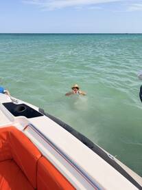 A joyful swimmer gives a thumbs up in the clear waters, with a boat in the foreground, capturing the essence of fun and relaxation on a sunny day.
