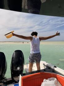 A person joyfully stands on the edge of a boat, arms outstretched, enjoying the beautiful beach scenery — a perfect moment of celebration on the water.