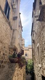 A narrow alleyway between stone buildings, featuring potted plants and a clear sky above. The image lacks clarity and focus.