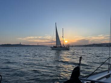 A picturesque sunset view of a sailing boat gliding through the waters of Lisbon, with a vibrant sky and iconic landmarks in the background — perfect for romantic evening cruises.