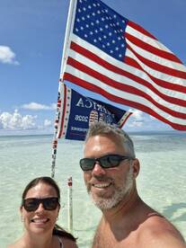 A couple poses for a selfie at the beach with a vibrant American flag in the background, capturing a moment of celebration and fun in the sun.