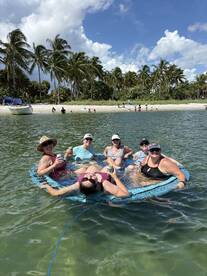A lively group enjoys a fun day on a floating mat in the water, surrounded by palm trees and a beautiful beach — perfect for summer gatherings and beach parties.