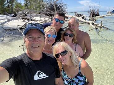A cheerful group of friends takes a selfie on a beautiful beach, surrounded by clear waters and driftwood — perfect for capturing memorable moments during a fun day out.