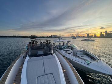 A lively group celebrates aboard two boats at sunset, with a stunning city skyline in the background — perfect for unforgettable boating experiences.