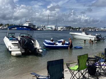 A lively group enjoys a day at the beach with boats anchored nearby, featuring swimming and relaxation on inflatable loungers — perfect for summer fun.