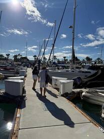 A couple strolls through a vibrant marina, surrounded by various boats and a clear blue sky — perfect for exploring the waterfront.